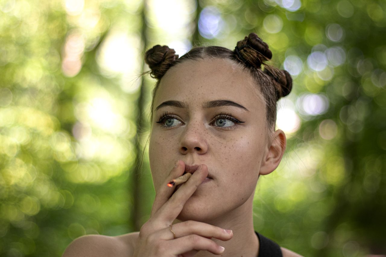 Young female with stylish hairdo smoking weed while standing against blurred background of green trees