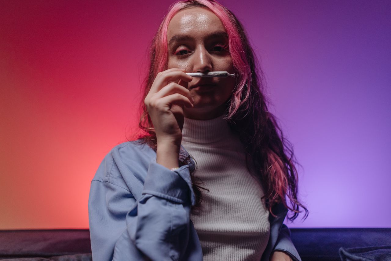 Woman with pink hair holding joint in studio with vibrant lighting. Cannabis themed.